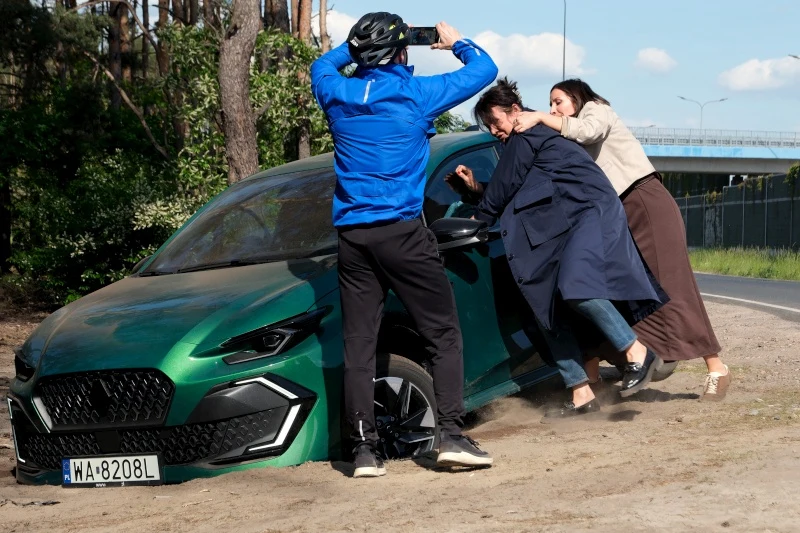 Szczęśliwie żadnej z kobiet nic się nie stanie Trzy osoby w dynamicznej interakcji przy zielonym samochodzie osobowym, z czego jedna w kasku rowerowym fotografuje sytuację telefonem, a dwie stoją przy drzwiach próbując otworzyć pojazd lub pomóc kierowcy.