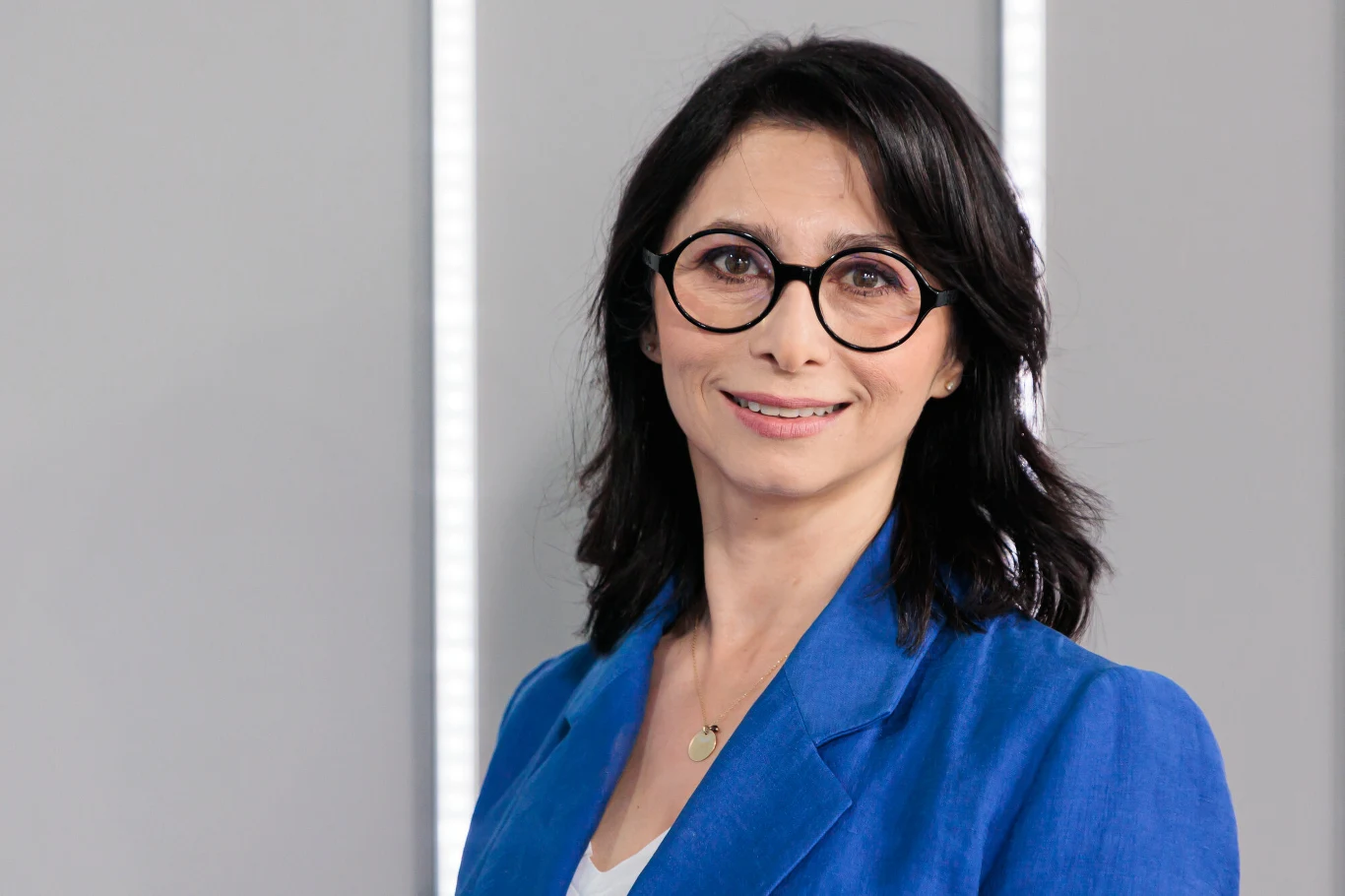 Kobieta o ciemnych, półdługich włosach i okrągłych okularach ubrana w niebieską marynarkę, uśmiecha się delikatnie, stojąc na tle jasnej ściany z pionowymi, jasnymi pasami.