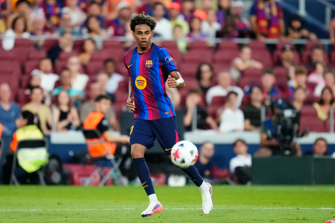 Lamine Yamal w barwach FC Barcelona 2026/2027 Piłkarz w stroju FC Barcelony prowadzi piłkę po murawie stadionu podczas meczu, w tle widoczni kibice na trybunach.