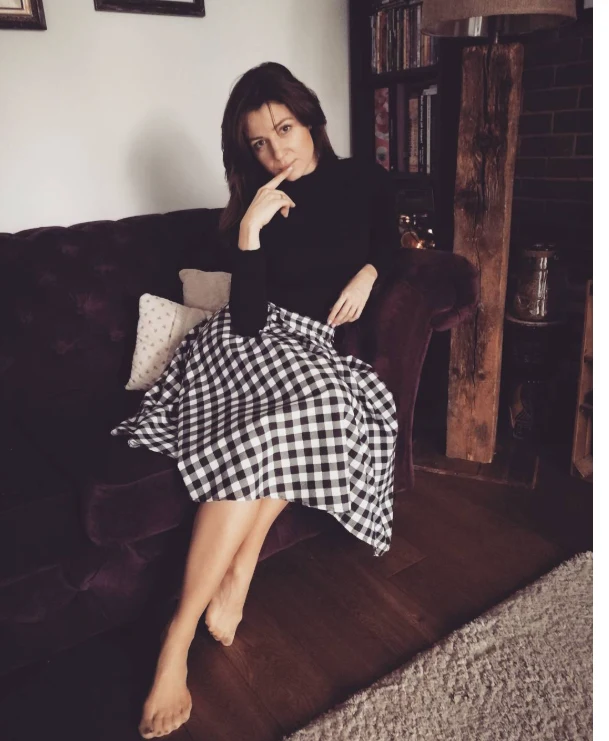 Aleksandra Zienkiewicz w salonie. Kobieta w czarnej bluzce i kraciastej spódnicy siedzi boso na ciemnej sofie, opierając się wygodnie i wspierając dłonią brodę, z półkami na książki i elementami drewnianymi w tle.