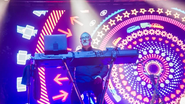Mężczyzna grający na syntezatorze na scenie koncertowej, otoczony kolorowymi, psychodelicznymi iluminacjami świetlnymi z motywami geometrycznymi i figurami w tle.