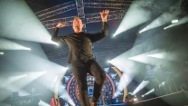 Wokalista w dynamicznej pozie na scenie, otoczony efektownym oświetleniem, z tyłu widoczny kolorowy, podświetlony ekran oraz instrumentaliści grający na klawiszach.