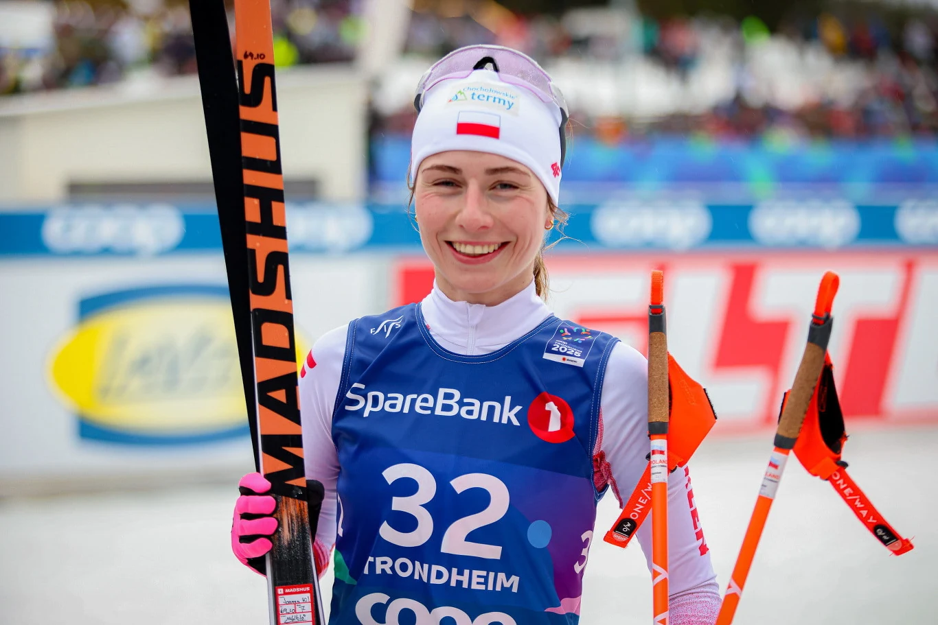 Joanna Kil Uśmiechnięta sportsmenka w stroju narciarskim trzyma w dłoniach parę nart oraz kijki, mając na sobie niebieską kamizelkę z numerem 32 i nazwami sponsorów.