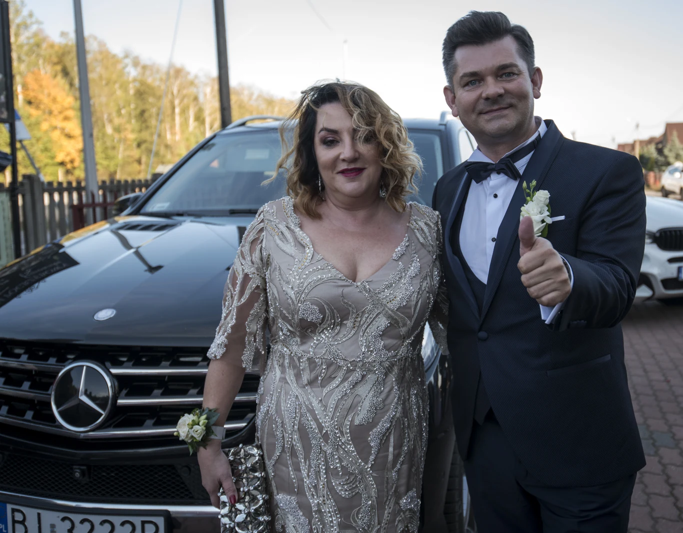 Elegancko ubrana kobieta w jasnej sukni i mężczyzna w garniturze z muszką stoją obok siebie, uśmiechnięty mężczyzna pokazuje uniesiony kciuk, za nimi czarny samochód marki Mercedes.
