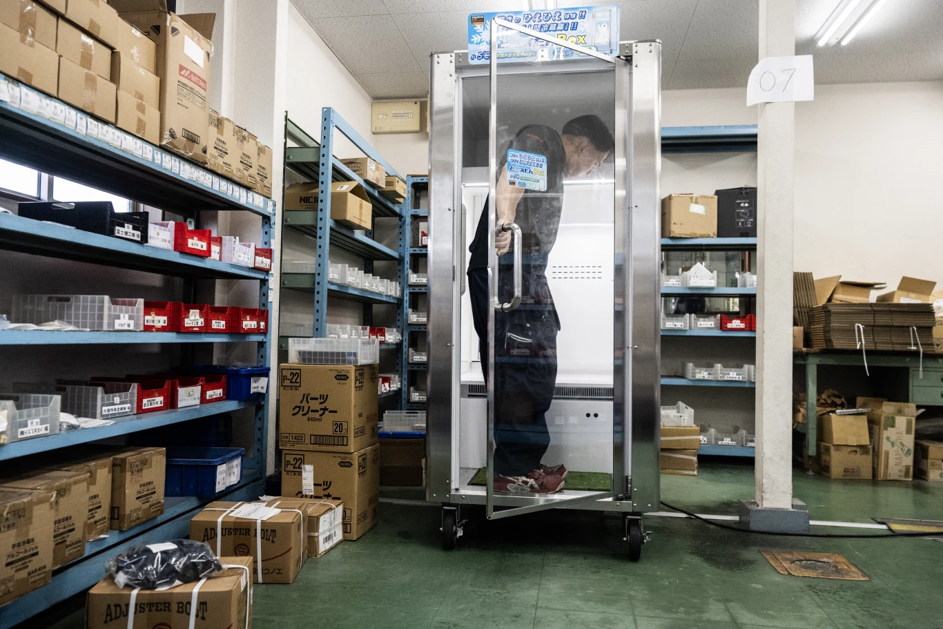 Mężczyzna zamknięty w przezroczystej kabinie, otoczony metalowymi regałami z kartonami i plastikowymi pojemnikami w magazynie.