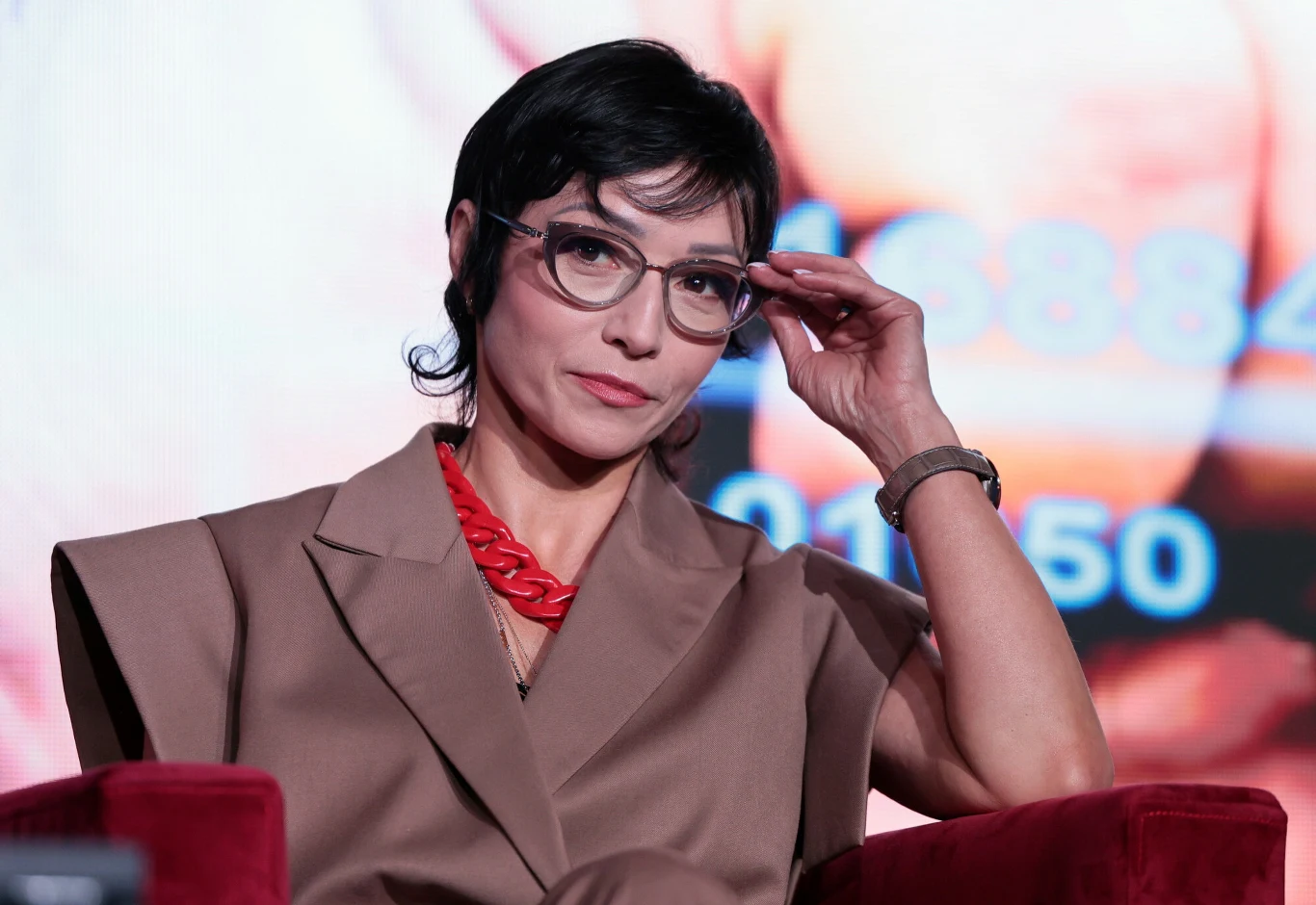 Barbara Mroz-Gorgon Barbara Mroz-Gorgon w okularach, brązowym garniturze i czerwonej biżuterii siedzi na czerwonym fotelu.