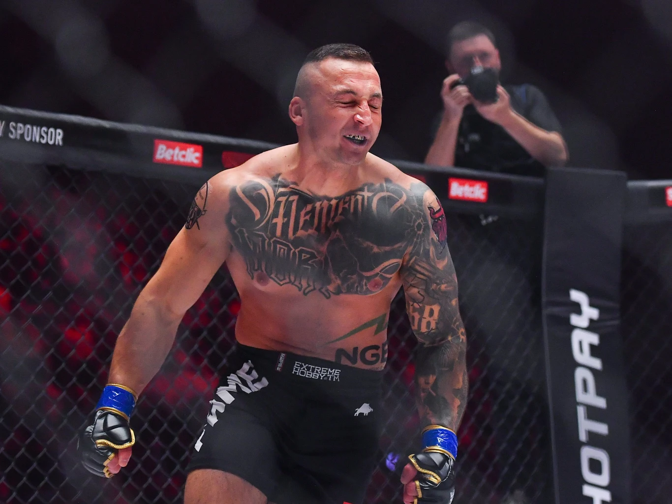 Bartosz Szachta zmierzy się z Kasjuszem Życińskim w walce wieczoru podczas gali FAME MM 32 Zawodnik MMA w czarnych spodenkach i z licznymi tatuażami na tułowiu oraz ramionach stoi w oktagonie z wyrazem emocji na twarzy, w tle fotograf wykonujący zdjęcia przez siatkę klatki.