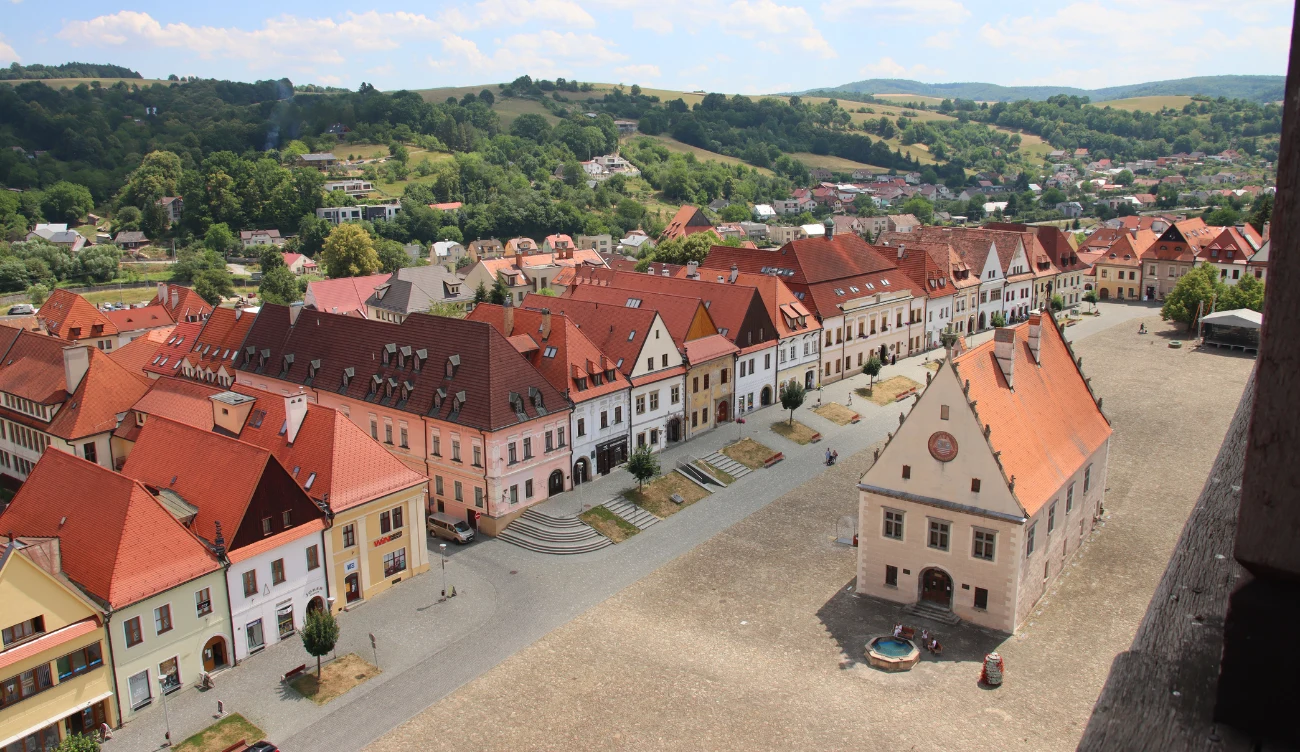 Jakie są średniowieczne mury obronne z basztą w Bardejowie? Średniowieczne mury obronne i baszta w Bardejowie, kolorowe kamienice, rynek z ratuszem, fontanną i wzgórza w tle.