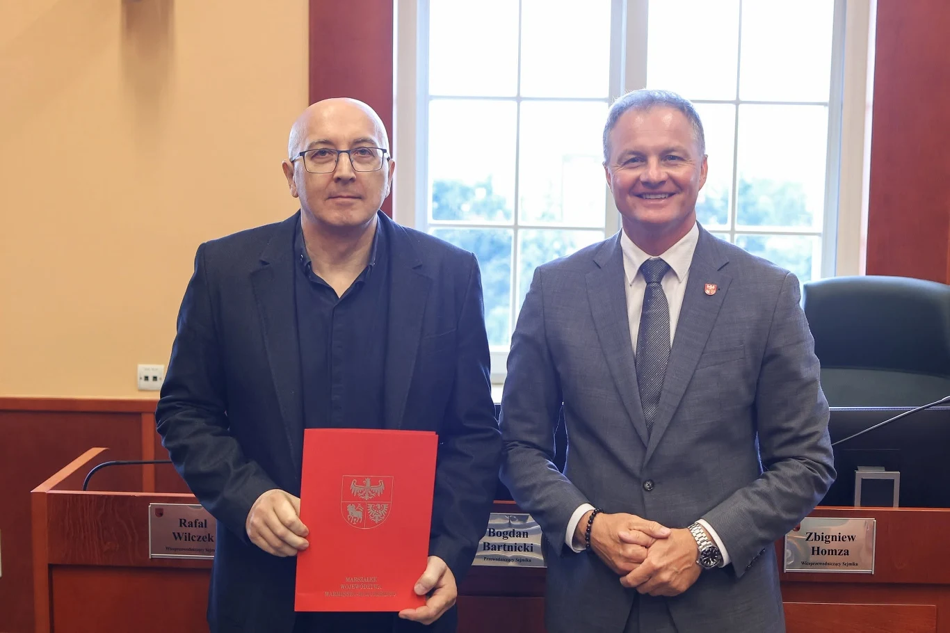 Robert Marzewski z AstroLab i marszałek województwa warmińsko-mazurskiego Marcin Kuchciński Robert Marzewski i Marcin Kuchciński stoją w sali obrad, jeden z nich trzyma czerwoną teczkę z polskim godłem.