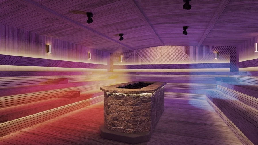 Green House Development Przestronna, nowoczesna sauna z drewnianymi ławkami rozmieszczonymi na kilku poziomach oraz dużym, kamiennym piecem na środku, podświetlona wielokolorowym światłem LED.