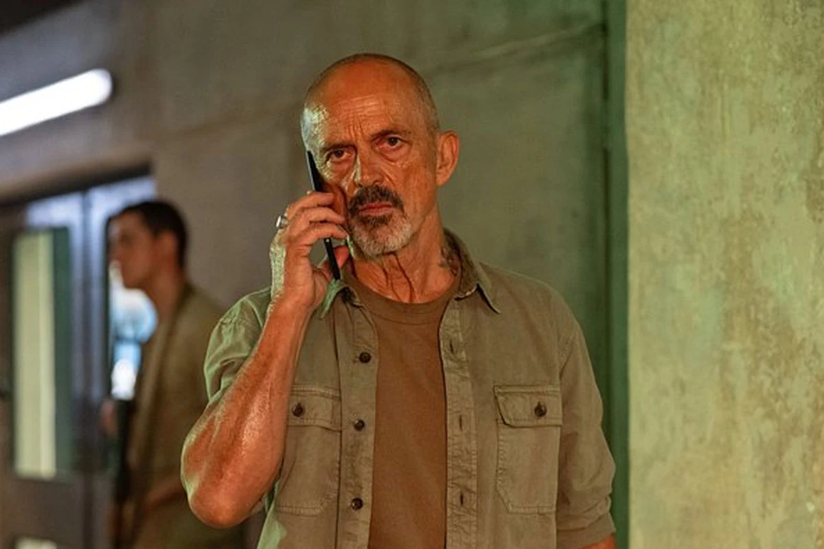 Jacek Koman w serialu "Apollo w ogniu: Apollo Has Fallen" Jacek Koman w serialu "Apollo w ogniu: Apollo Has Fallen", stoi przy betonowej ścianie, rozmawia przez telefon.