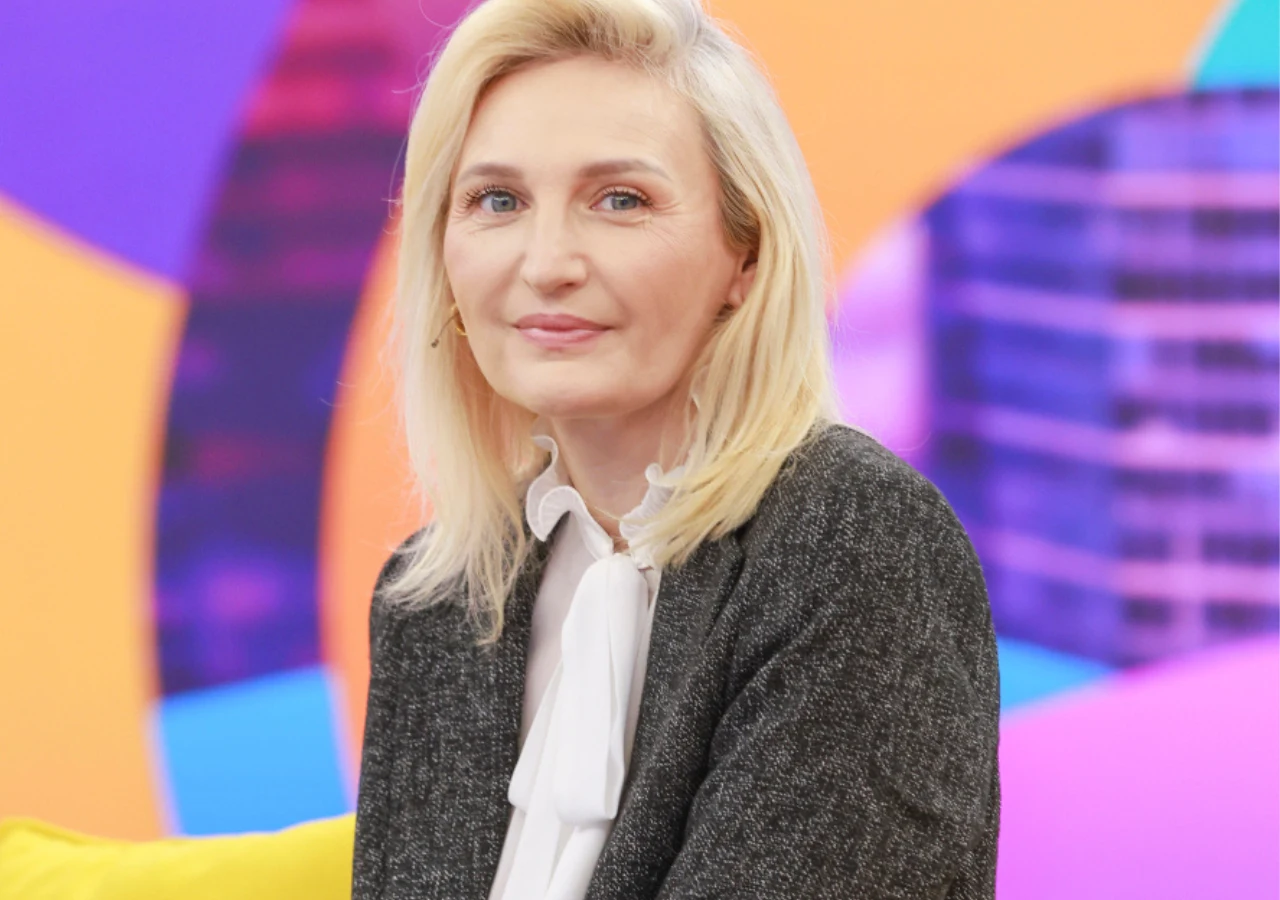 Kobieta o blond włosach w eleganckiej, ciemnej marynarce i białej koszuli z kokardą siedzi na żółtej sofie, na tle kolorowej abstrakcyjnej grafiki.