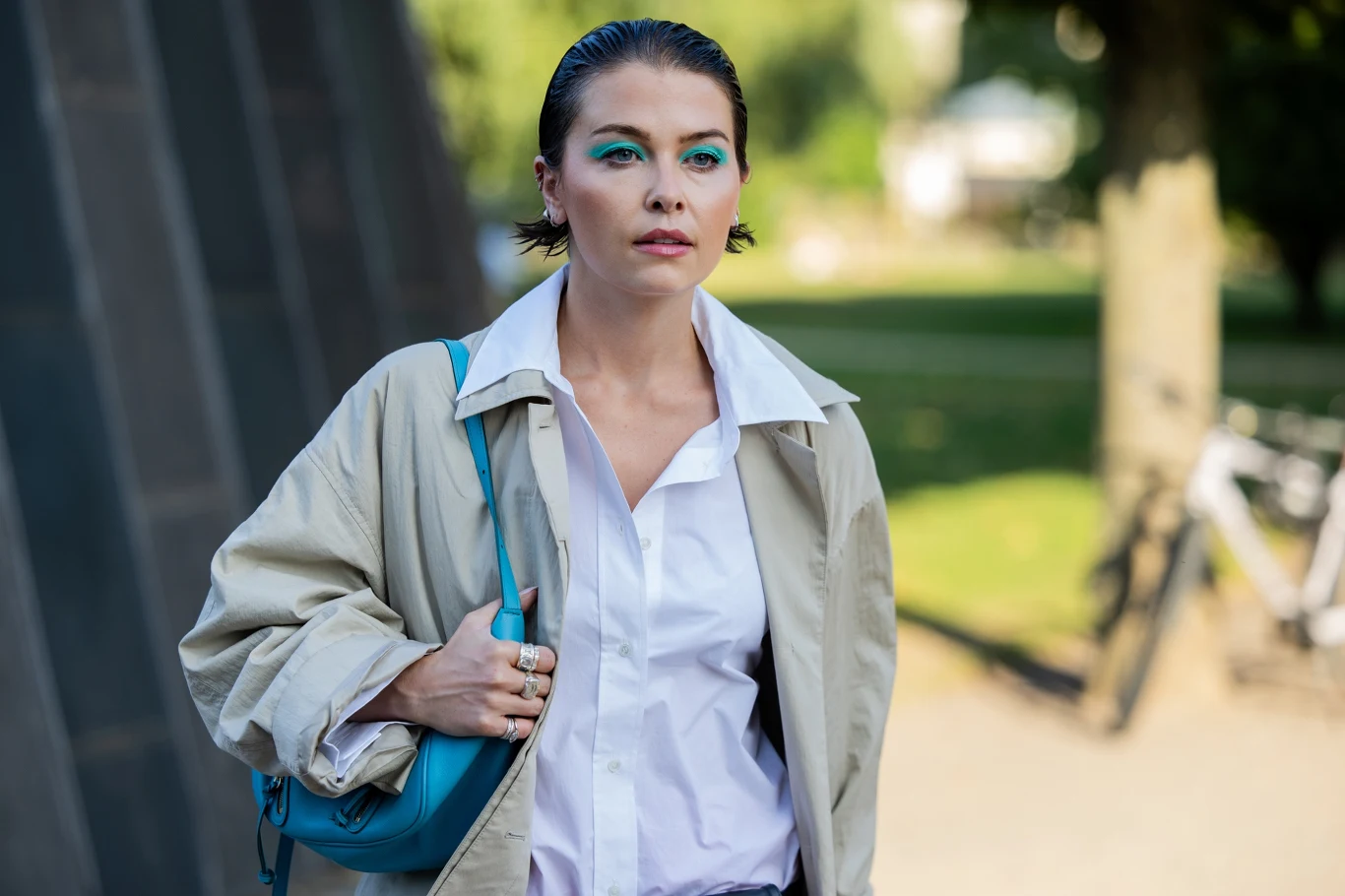 Jesienią aqua blue rozświetli stylizacje. Ten kolor pokochały już it-girls Kobieta z makijażem oczu w kolorze niebieskim, w białej koszuli i beżowej kurtce, z niebieską torebką na ramieniu.