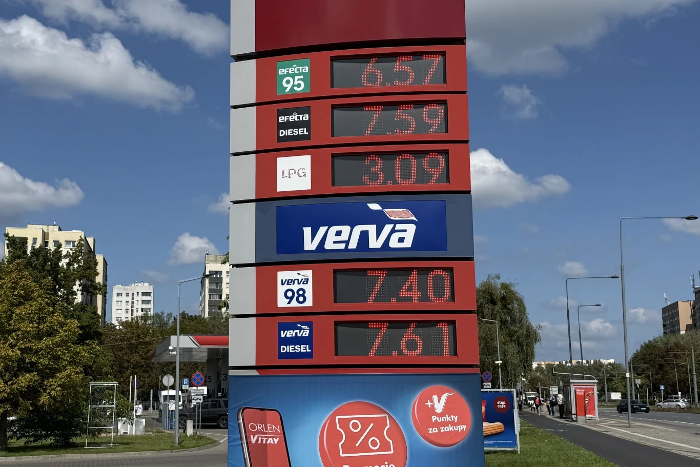 Ceny paliw na stacji Orlen 25 sierpnia 2026. Stacja paliw Orlen z cyfrowym wyświetlaczem cen paliw: benzyna 95, diesel, LPG, Verva 98 i Verva Diesel, na tle miejskiej ulicy, budynków mieszkalnych i drzew.