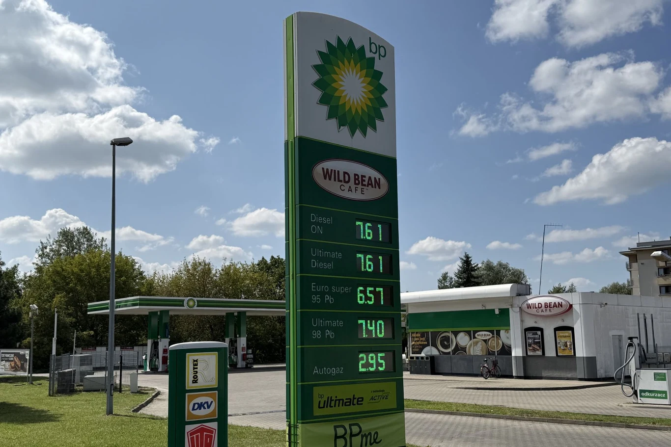 Ceny paliw na BP. Stacja paliw BP z charakterystycznym słupem cenowym, na którym widoczne są aktualne ceny różnych rodzajów paliwa, w tle budynek stacji oraz logo kawiarni Wild Bean Café.