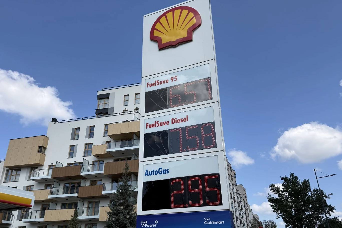 Ceny paliw na Shell. Tablica cenowa stacji paliw Shell z widocznymi cenami dla benzyny, diesla i autogazu na tle nowoczesnych budynków mieszkalnych oraz nieba z chmurami.