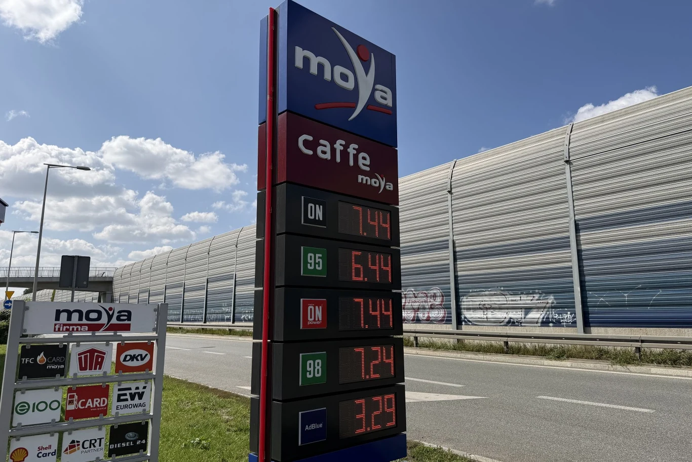 Ceny paliw na stacji Moya. Słupek cenowy stacji paliw Moya z wyraźnie widocznymi cenami różnych rodzajów paliwa ustawiony przy ulicy, w tle ekrany akustyczne i fragment jezdni.