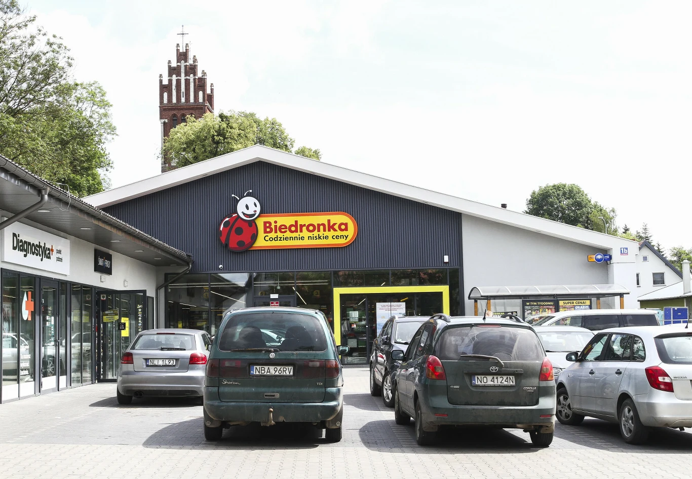 Czy można zostawić auto na noc na parkingu pod Biedronką lub Lidlem? Wejście do Biedronki, samochody zaparkowane na parkingu, w tle kościół z czerwoną wieżą