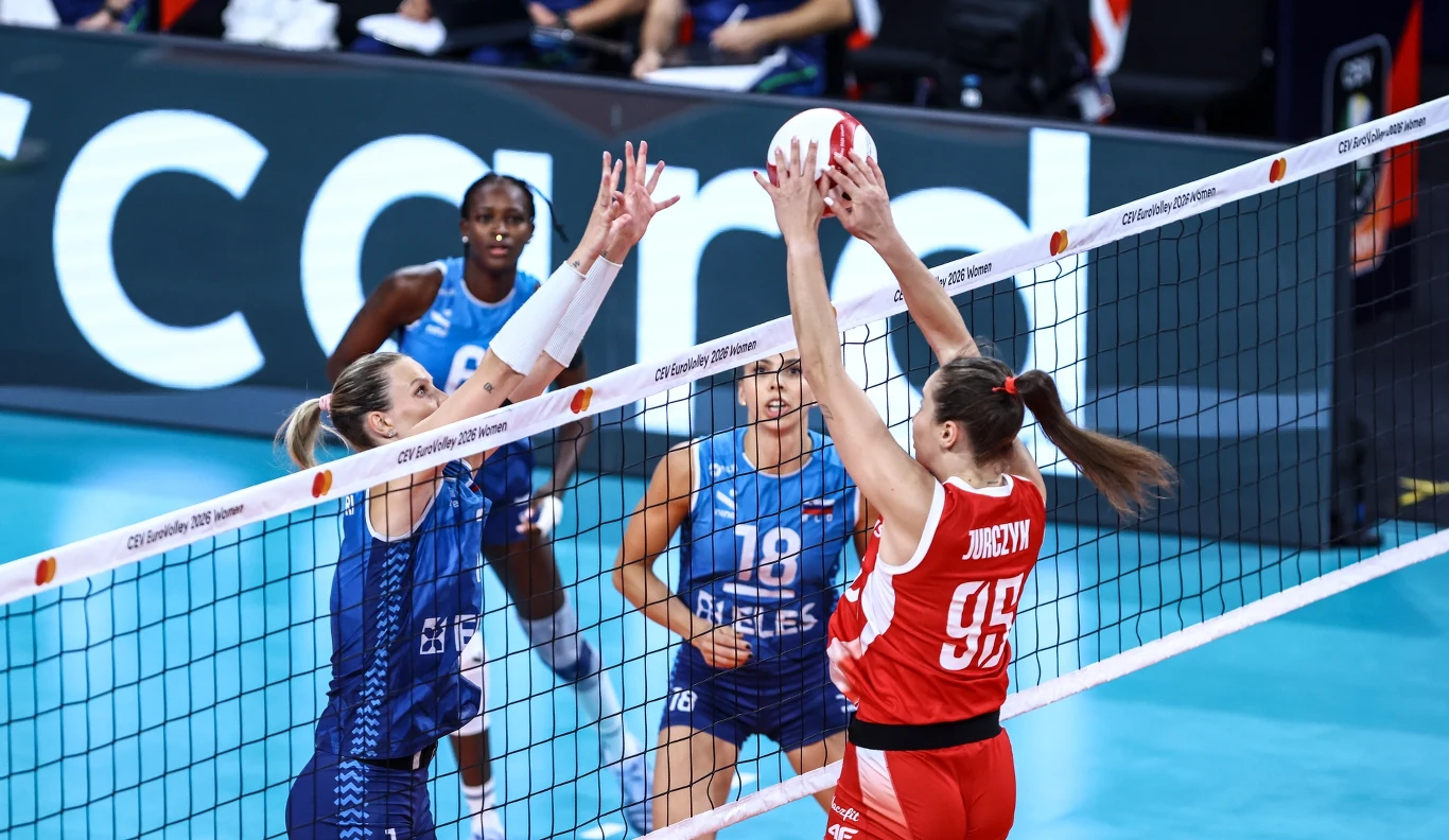 Mecz Polska - Słowenia, pod siatką walczy Magdalena Jurczyk Siatkarka w czerwonym stroju próbuje przebić piłkę przez siatkę, naprzeciw której dwie zawodniczki w niebieskich strojach skaczą do bloku, a kolejna siatkarka w tle jest gotowa do dalszej akcji.
