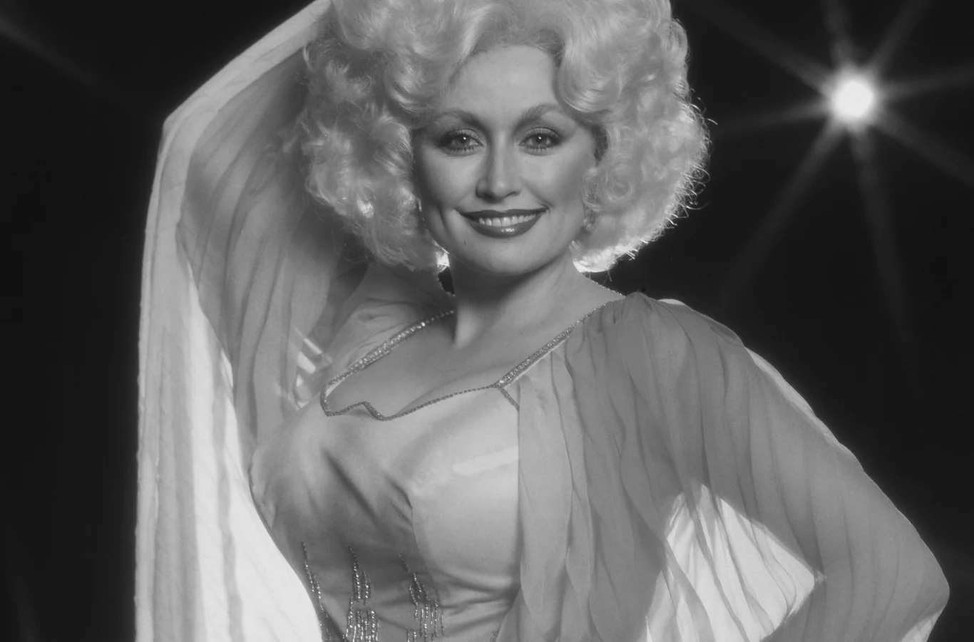 Dolly Parton (19 stycznia 1946 - 25 sierpnia 2026) Dolly Parton w eleganckiej sukni z szerokimi rękawami, jasne bujne włosy, ciemne tło z rozbłyskami światła.