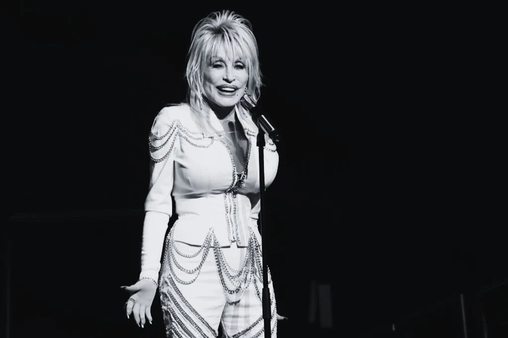 Donald Trump pożegnał piosenkarkę Dolly Parton Dolly Parton w ozdobnym stroju z frędzlami, stojąca przy mikrofonie na scenie, zdjęcie czarno-białe.
