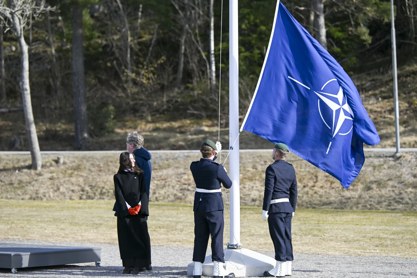 Dwóch żołnierzy stoi przy maszcie z powiewającą flagą NATO podczas ceremonii w bazie marynarki wojennej Musko. Obok stoją przedstawiciele szwedzkiego rządu.
