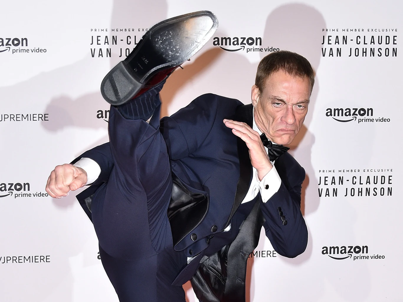Mężczyzna w eleganckim garniturze wykonuje wysokie kopnięcie, prezentując sprawność fizyczną na tle ściany z logotypami Amazon Prime Video i nazwą serialu Jean-Claude Van Johnson.