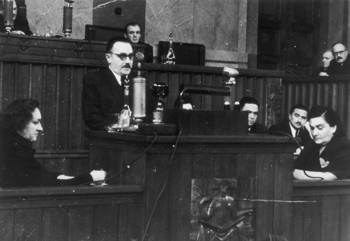 Bolesław Bierut podczas przemówienia w 1948 roku Sędzia w okularach prowadzi rozprawę sądową, przemawiając zza wysokiej ławy, a po bokach słucha go grupa skupionych osób ubranych na czarno.