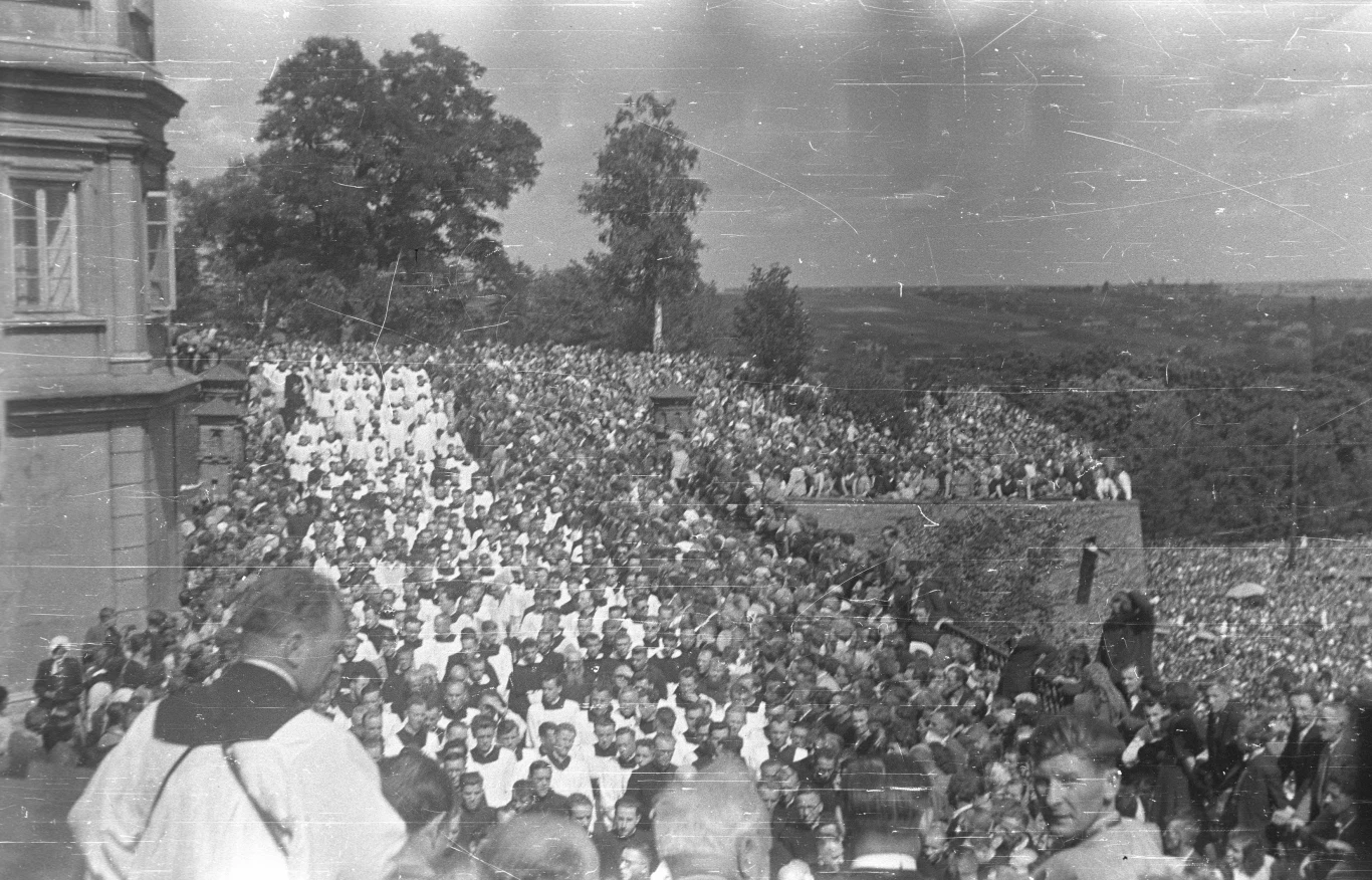 Jasna Góra podczas składania ślubów w 1956 roku Tłum ludzi zgromadzonych na zewnętrznych schodach prowadzących do dużego budynku, wśród których widoczni są duchowni oraz liczne osoby w ciemnych i jasnych ubraniach, otoczonych zielenią i otwartą przestrzenią.
