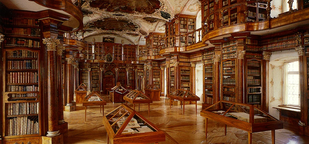 Bogato zdobione wnętrze biblioteki z wysokimi drewnianymi regałami pełnymi starych książek, podświetlonymi witrynami wypełnionymi manuskryptami i rękopisami na tle zabytkowego sufitu z malowidłami.