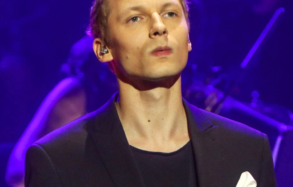 Igor Herbut: Mężczyzna w garniturze z białą poszetką stoi na scenie w teatralnym oświetleniu, w uchu ma mikrofon douszny.