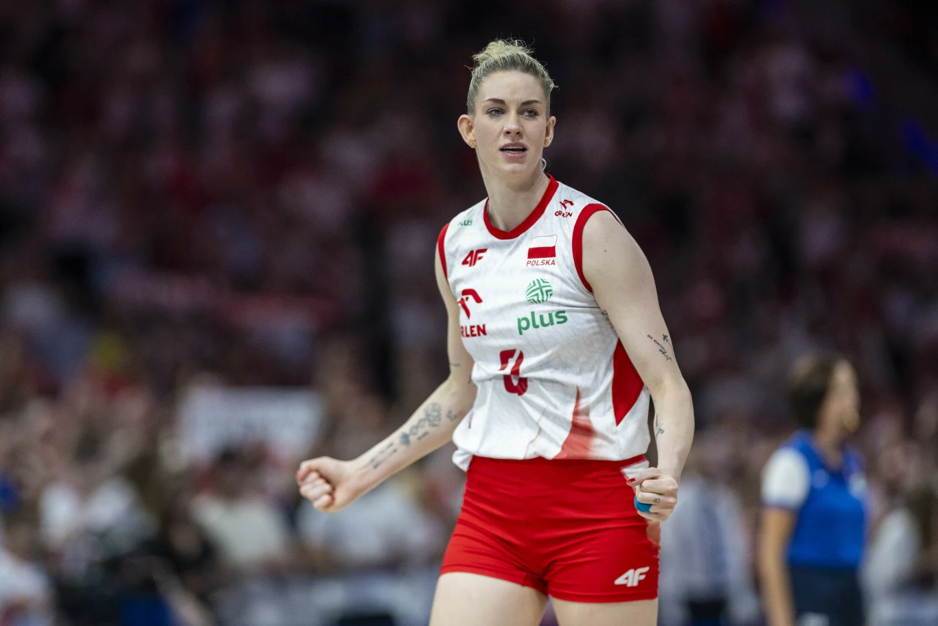 Magdalena Stysiak Siatkarka w biało-czerwonym stroju reprezentacji Polski wykonująca gest triumfu podczas meczu, otoczona rozmytym tłumem kibiców na trybunach.