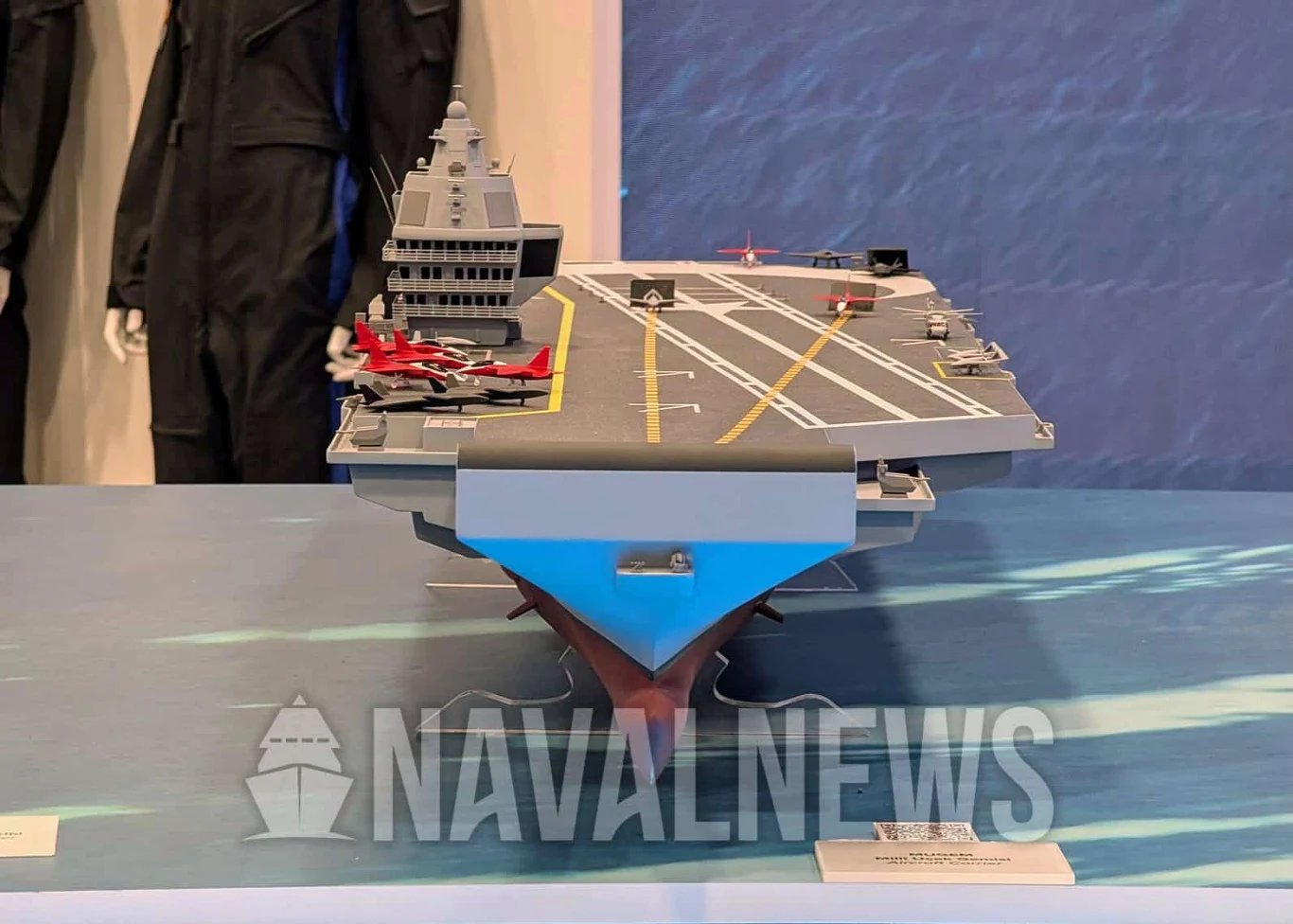 Makieta lotniskowca MUGEM. Model lotniskowca z żółtymi liniami na pokładzie startowym, kilkoma miniaturowymi samolotami odrzutowymi i śmigłowcem, z mostkiem dowodzenia po lewej stronie oraz ekspozycją mundurów w tle.