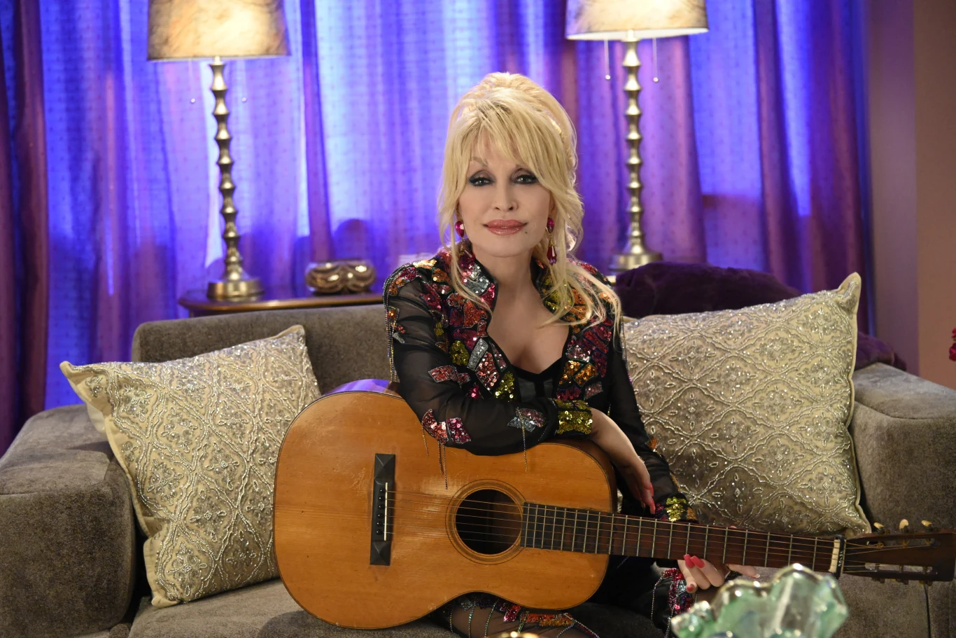 Dolly Parton to artystka, która nie bała się być "jakaś" w dobie podążania za trendami Kobieta z blond włosami siedzi na sofie, trzymając gitarę na kolanach, otoczona ozdobnymi poduszkami i dwiema lampami stojącymi po bokach.