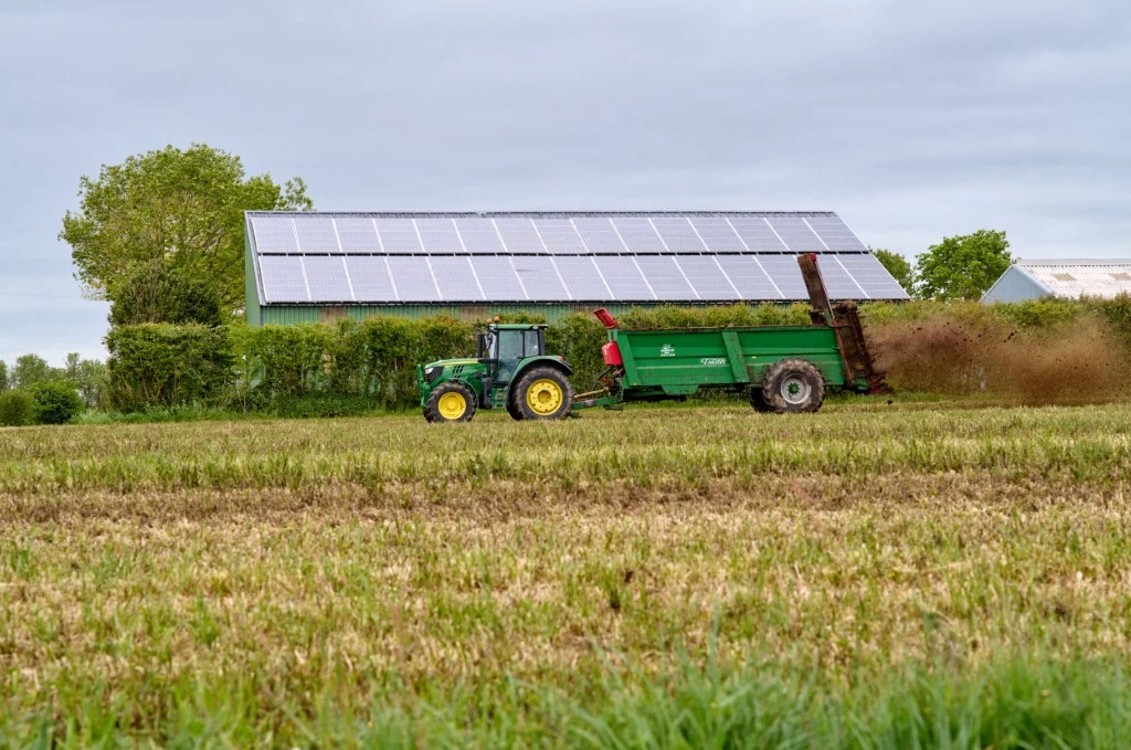 Agrowoltaika to jednoczesne wykorzystanie tego samego obszaru ziemi do upraw rolnych i produkcji energii elektrycznej z paneli słonecznych Traktor z zieloną przyczepą nawozi pole, w tle stodoła z panelami fotowoltaicznymi na dachu.