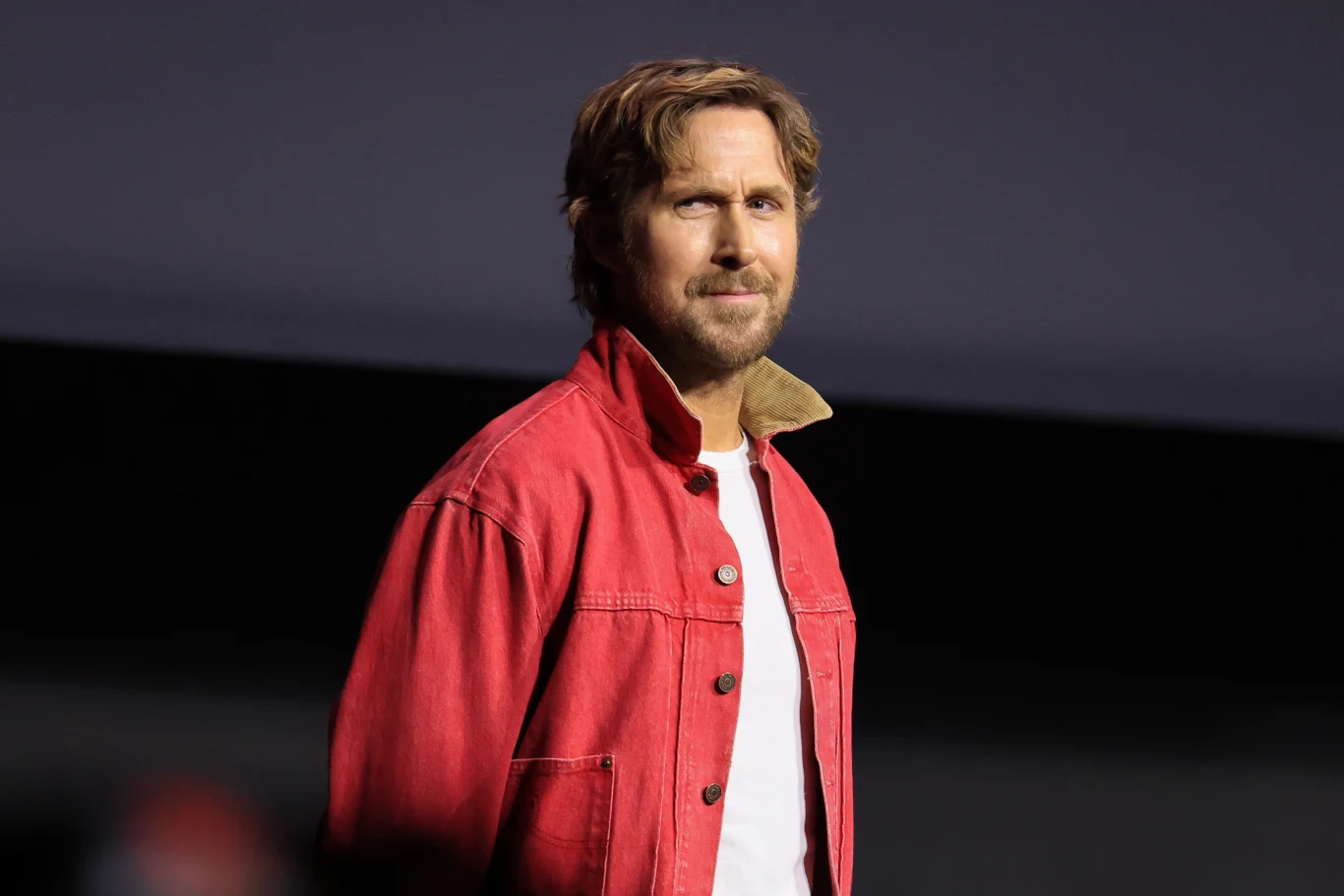 Ryan Gosling w lipcu 2026 roku Ryan Gosling w czerwonej kurtce, z brodą i średniej długości włosami, lipiec 2026, ciemne tło.