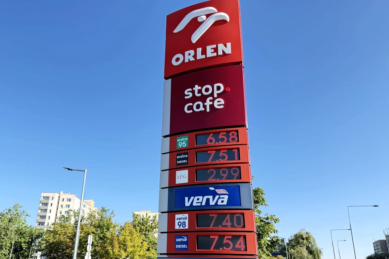 Ceny paliw na Orlenie. Cennik stacji paliw Orlen z aktualnymi cenami benzyny, oleju napędowego, LPG i paliw premium na wysokim pylonie, na tle bezchmurnego nieba oraz fragmentów zabudowy miejskiej i zieleni.