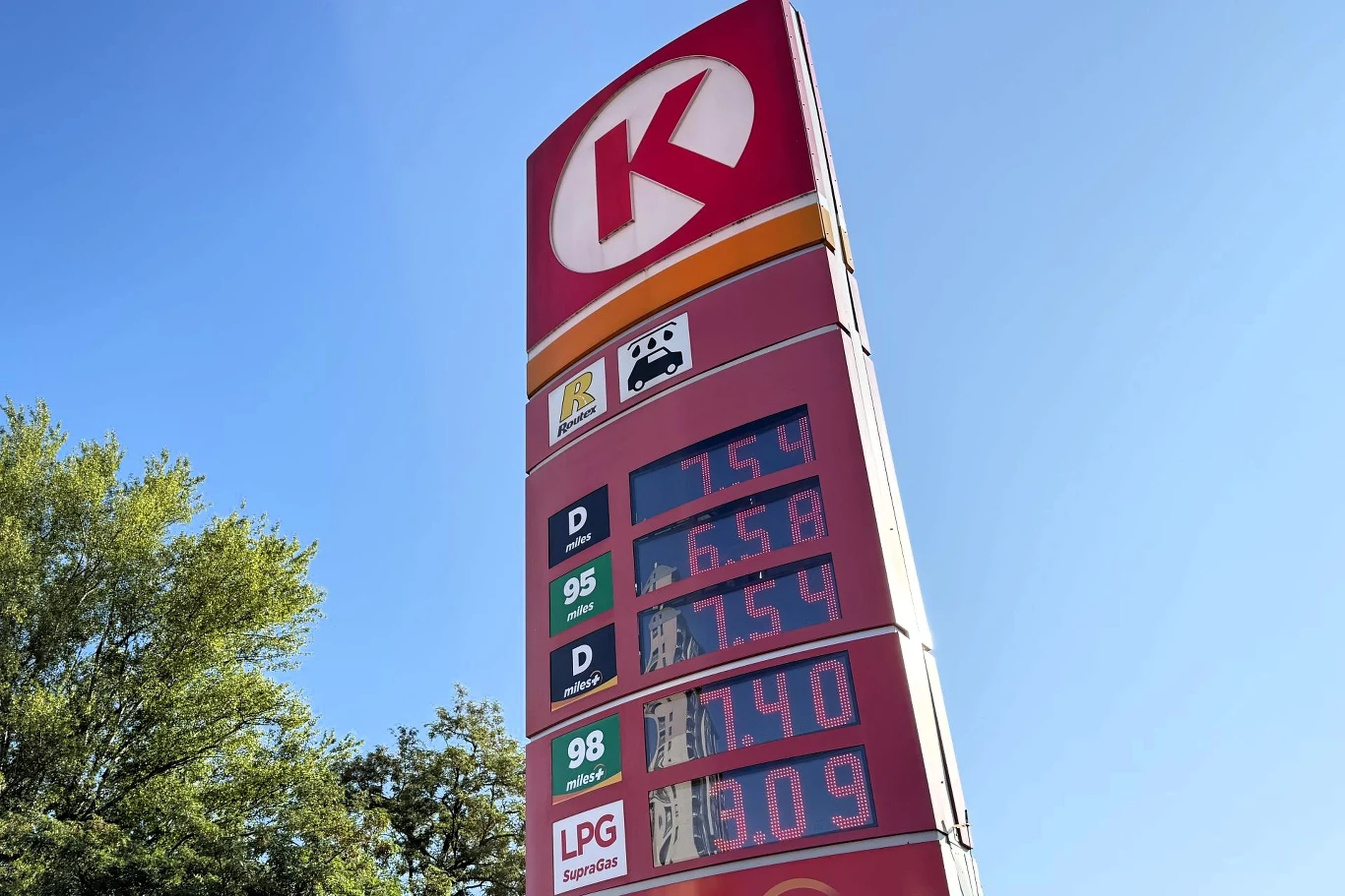 Ceny paliw na Circle K. Tablica cenowa stacji paliw Circle K z wyświetlonymi aktualnymi cenami różnych rodzajów paliwa na tle błękitnego nieba i koron drzew.