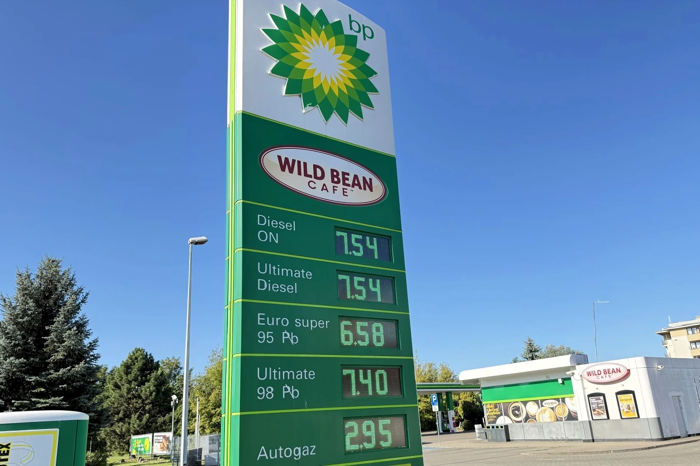 Ceny paliw na BP. Zielona tablica informacyjna stacji BP z wyświetlonymi cenami paliw, w tle widoczny budynek Wild Bean Cafe oraz dystrybutory paliwa na stacji benzynowej.