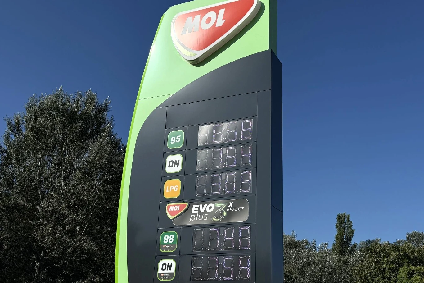 Ceny paliw na stacji MOL. Stacja paliw MOL z dużym, zielonym pylonem wyświetlającym aktualne ceny różnych rodzajów paliwa na tle drzew i błękitnego nieba.