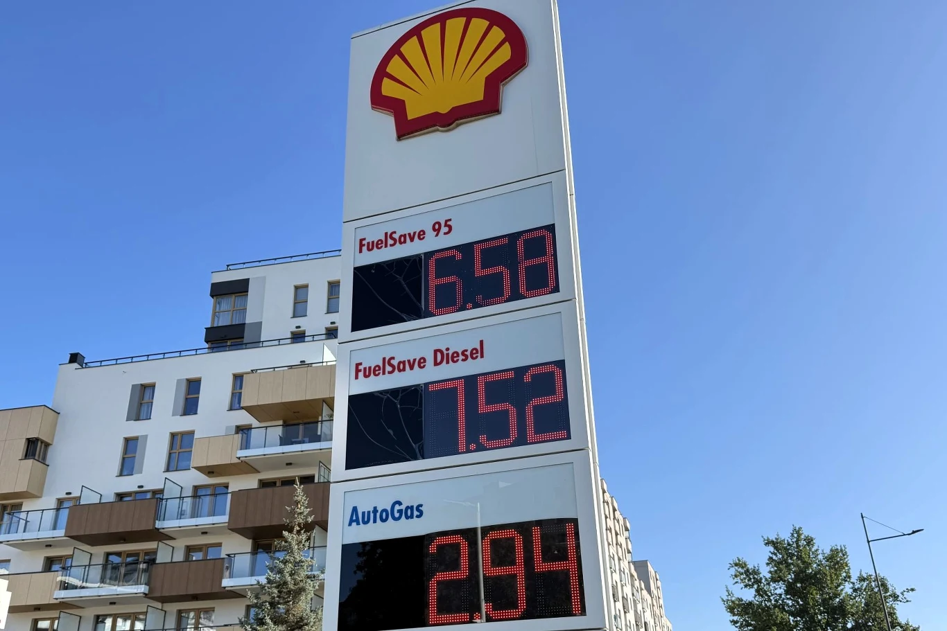 Ceny paliw na Shell. Tablica cenowa na stacji paliw Shell z aktualnymi cenami benzyny, diesla i autogazu, w tle nowoczesny budynek mieszkalny oraz częściowo widoczne drzewa i fragment nieba.