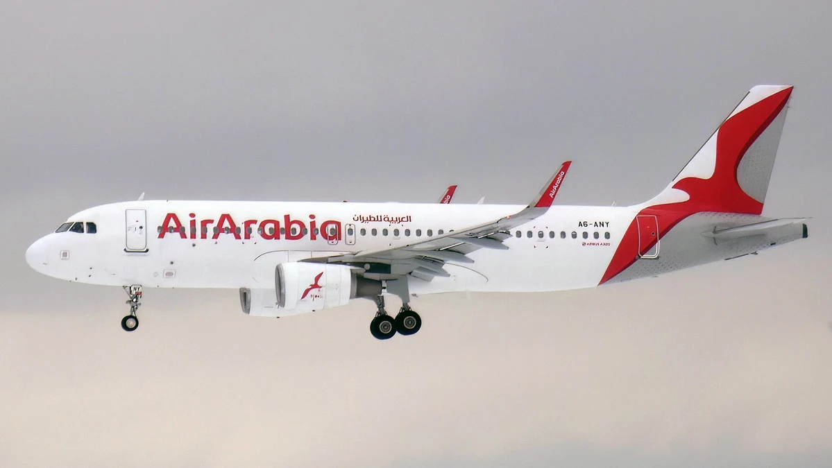 Samolot pasażerski linii Air Arabia w biało-czerwonym malowaniu z wysuniętym podwoziem podczas lotu na tle jasnego, jednolitego nieba.