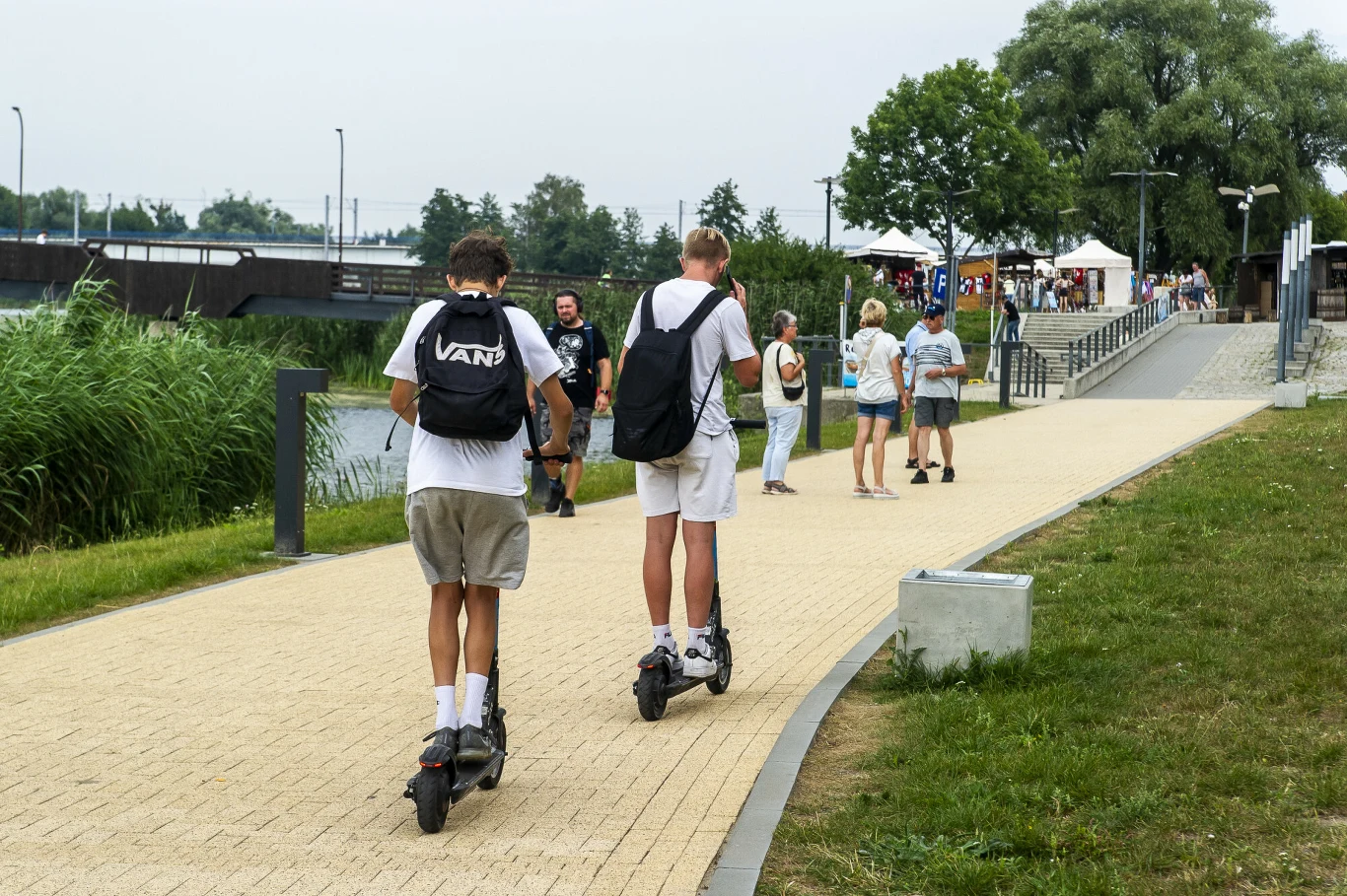 Obowiązek zakładania kasku niewiele zmienił Dwóch młodych mężczyzn na elektrycznych hulajnogach jedzie ścieżką w kierunku tłumu spacerowiczów, w tle zielona roślinność i biała zabudowa gastronomiczna.