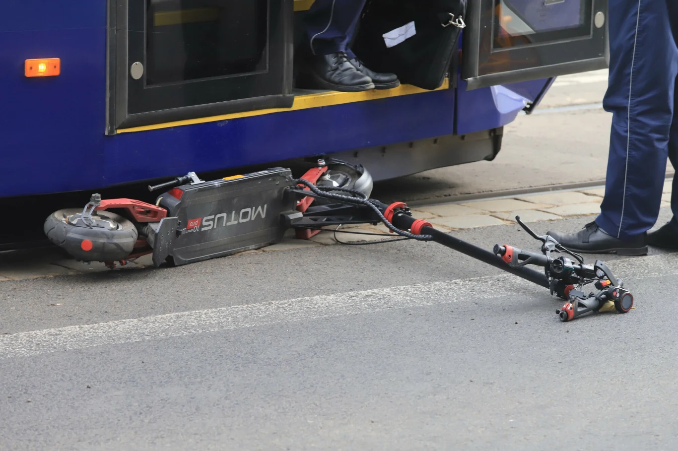 Statystyki wypadków z udziałem poruszających się hulajnogami małoletnich są porażające Elektryczna hulajnoga przewrócona i częściowo przygnieciona pod autobusem miejskim, obok stoją nogi dwóch osób w ciemnych spodniach.