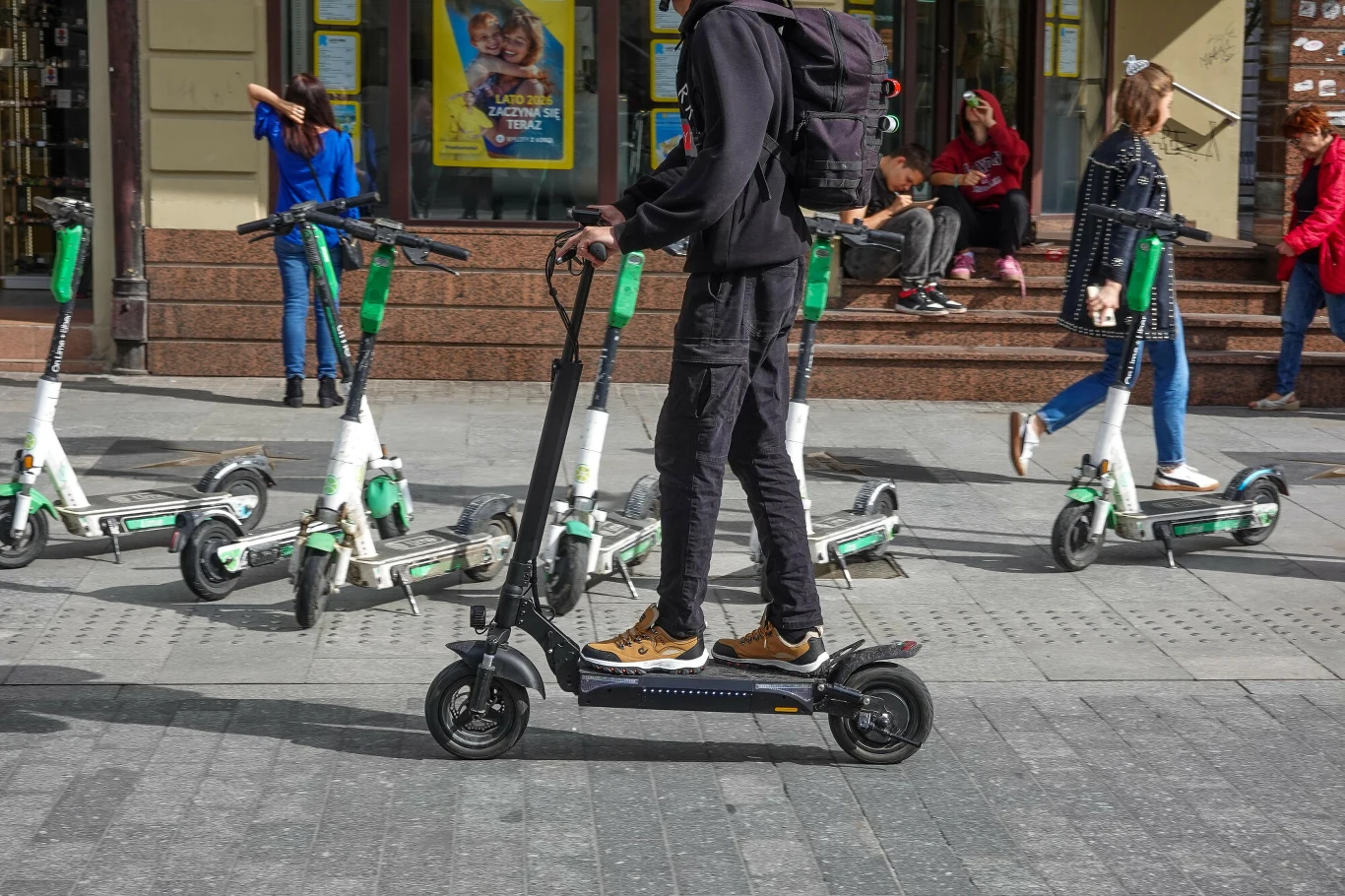 Coraz więcej poważnych wypadków z udziałem użytkowników hulajnóg elektrycznych. Wiele z nich dotyczy małoletnich Mężczyzna na hulajnodze elektrycznej jedzie ulicą, w tle rząd hulajnóg i piesi na chodniku.