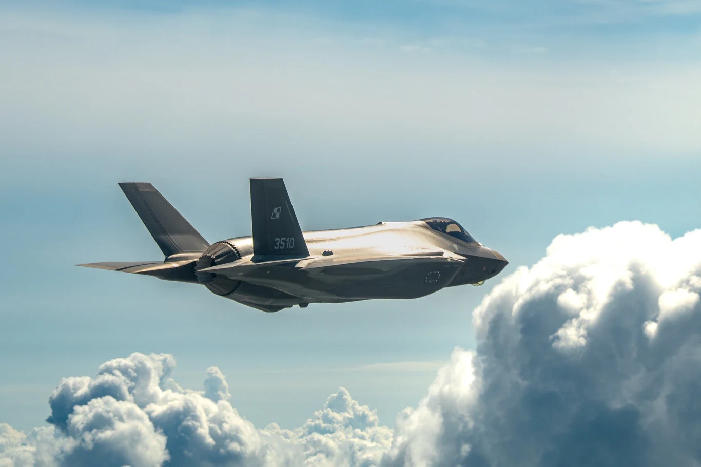 Pięć nowych F-35 leci do Polski. Skompletują połowę eskadry. Samolot F-35 z polską szachownicą w locie na dużej wysokości nad chmurami, widok z boku.