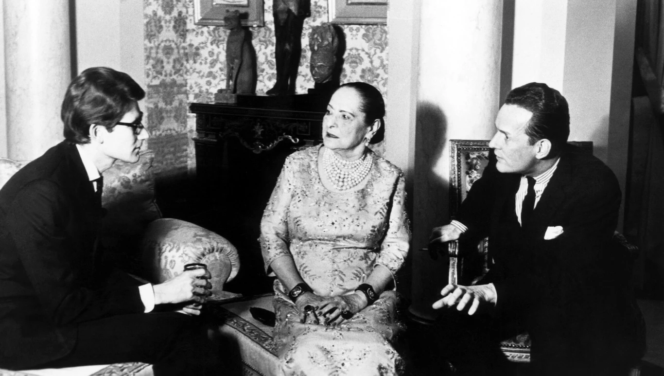 N zdjęciu: Yves Saint Laurent i Helena Rubinstein Trzy osoby w eleganckich strojach prowadzą poważną rozmowę w stylowo urządzonym wnętrzu, siedząc blisko siebie na fotelach i kanapie.