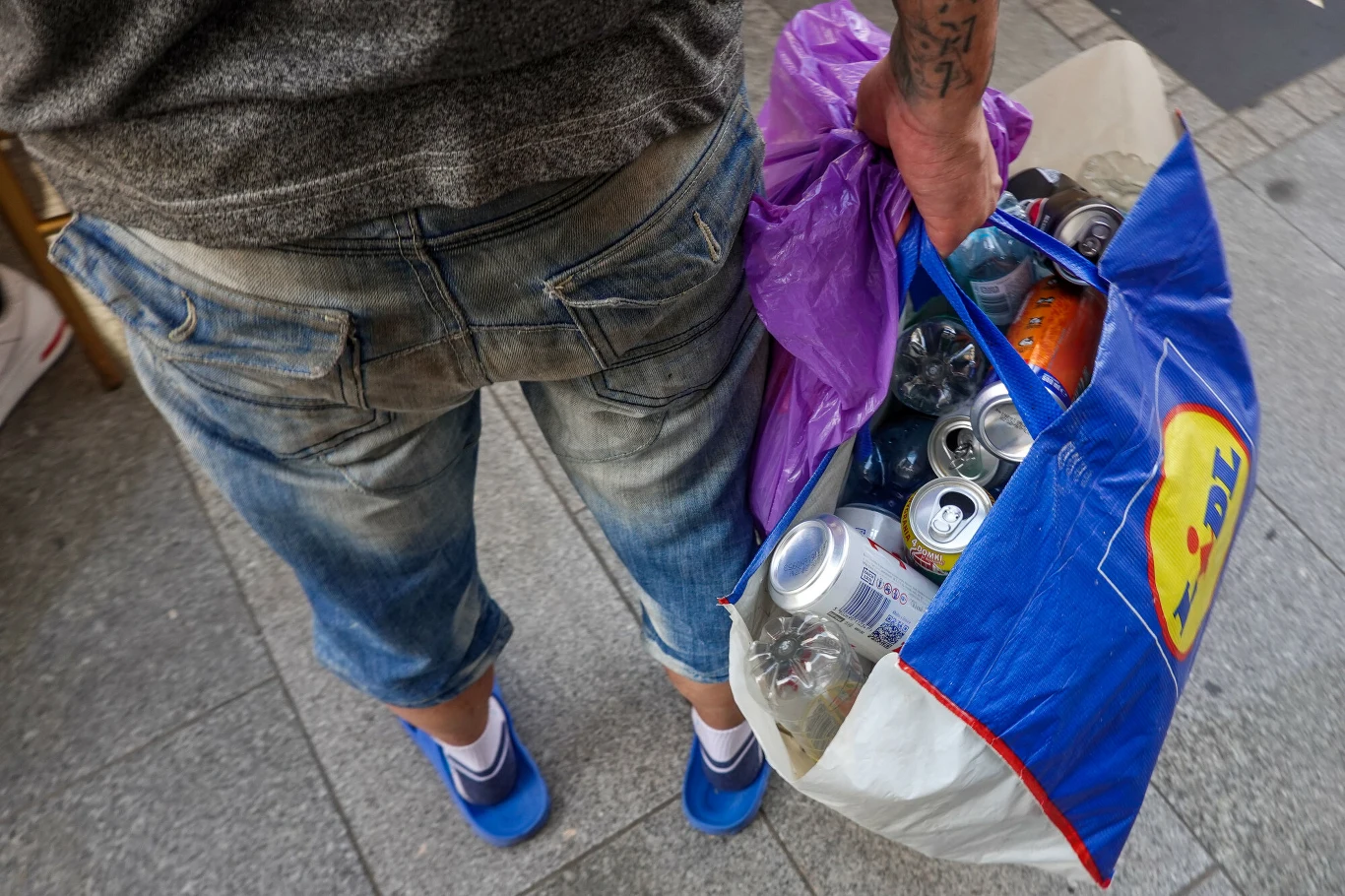 Aż 89 proc. ankietowanych zwraca opakowania w systemie kaucyjnym Mężczyzna w dżinsowych spodenkach i niebieskich butach trzyma torbę Lidl z pustymi puszkami i butelkami.