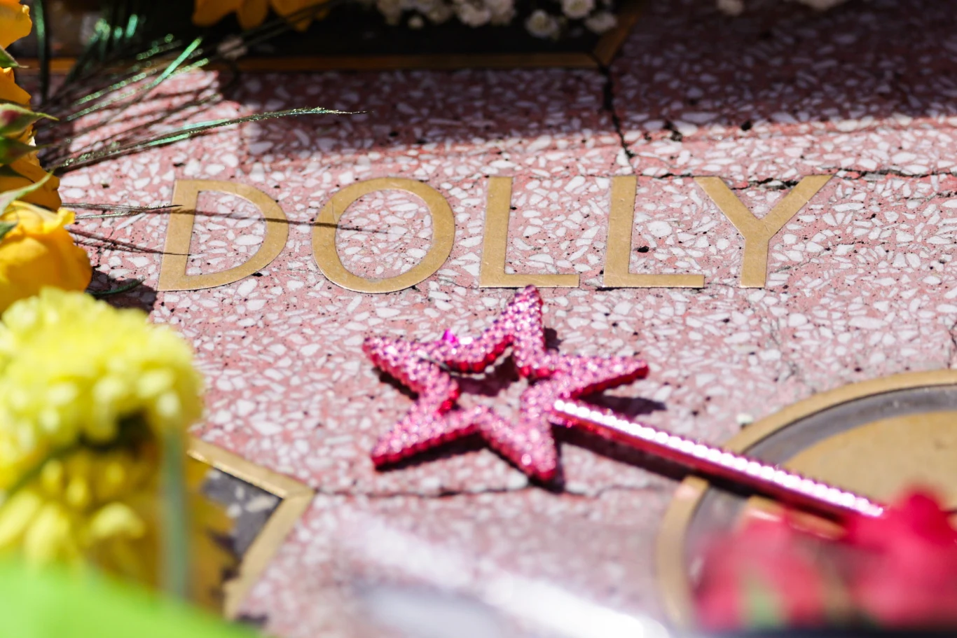 Gwiazda Dolly Parton w Alei Sław po jej śmierci Złoty napis DOLLY na jasnoróżowym marmurowym tle otoczonym żółtymi kwiatami i różdżką z różową gwiazdką.