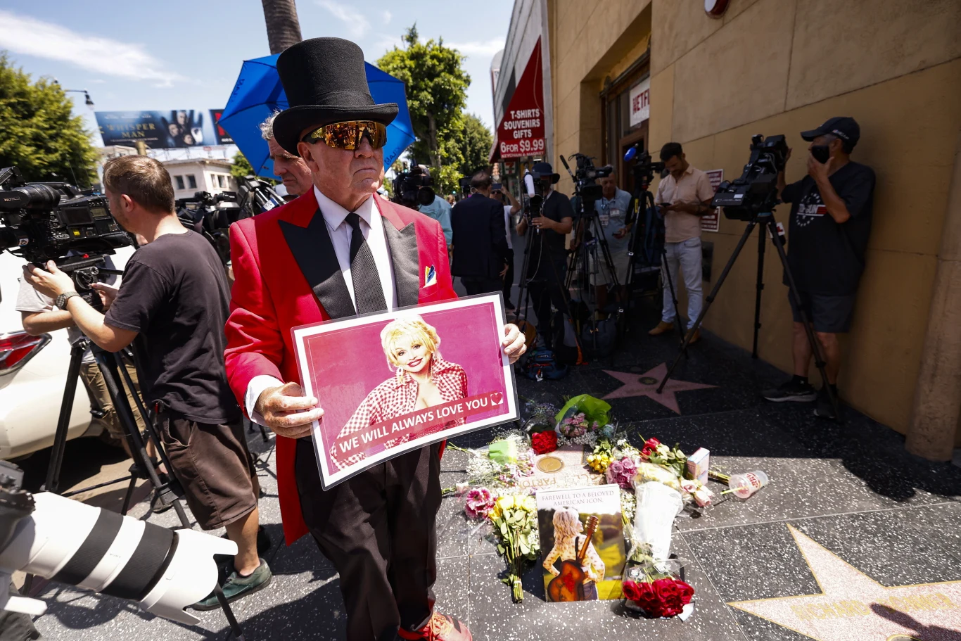 Fani upamiętniają Dolly Parton na Alei Sław w Los Angeles Mężczyzna w eleganckim stroju i cylindrze trzyma różowy plakat ze zdjęciem kobiety oraz napisem 'We will always love you' obok gwiazdy na Hollywood Walk of Fame, otoczonej kwiatami, świecami i pamiątkami; w tle kamery i reporterzy.